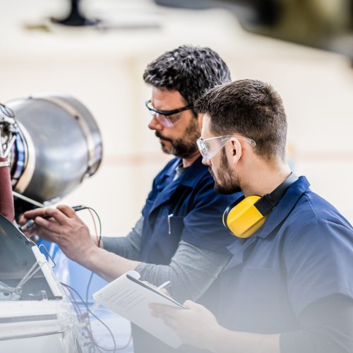 Aviation mechanic teaching coworker examining engine, side view. Aircraft hangar workers in protective equipment holding clipboard, medium shot